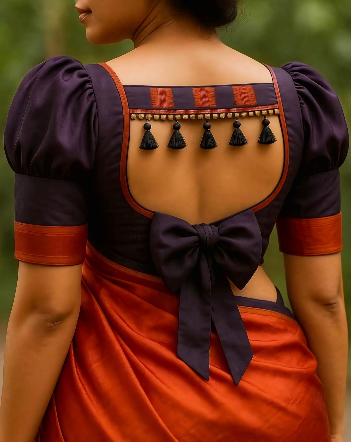 Purple and rust orange blouse with puff sleeves, square-cut back, tassel detailing, and large bow tie — stylish saree blouse design for festive occasions.