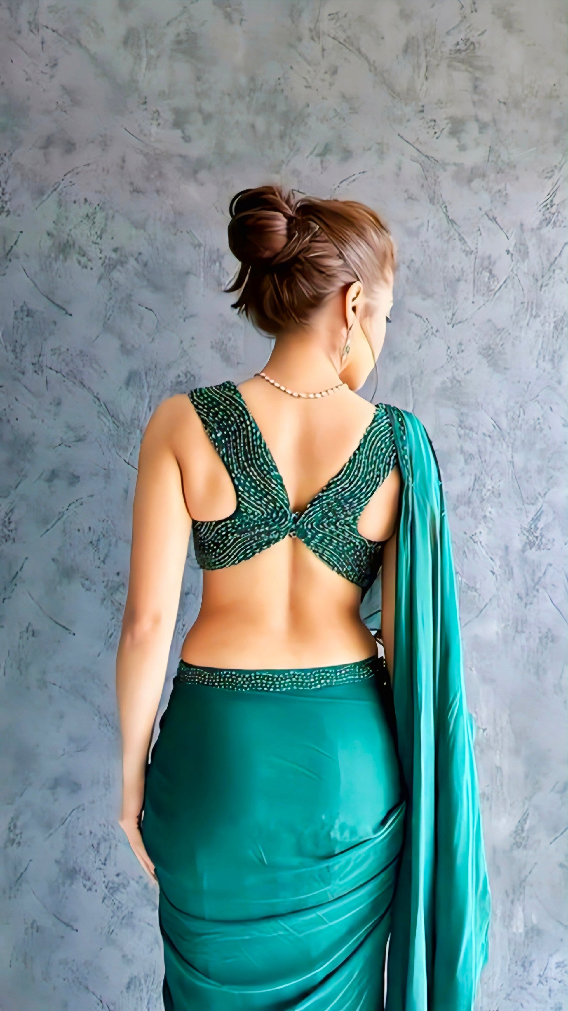 A woman wearing a green saree with a stylish twisted back-knot blouse, standing against a textured grey background.
