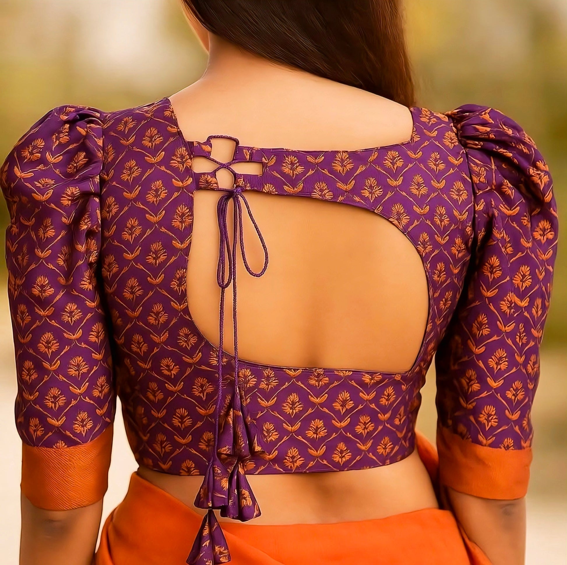 Woman wearing a purple brocade saree blouse with puff sleeves, square back cut, and dori tie-up — traditional silk ethnic blouse design.