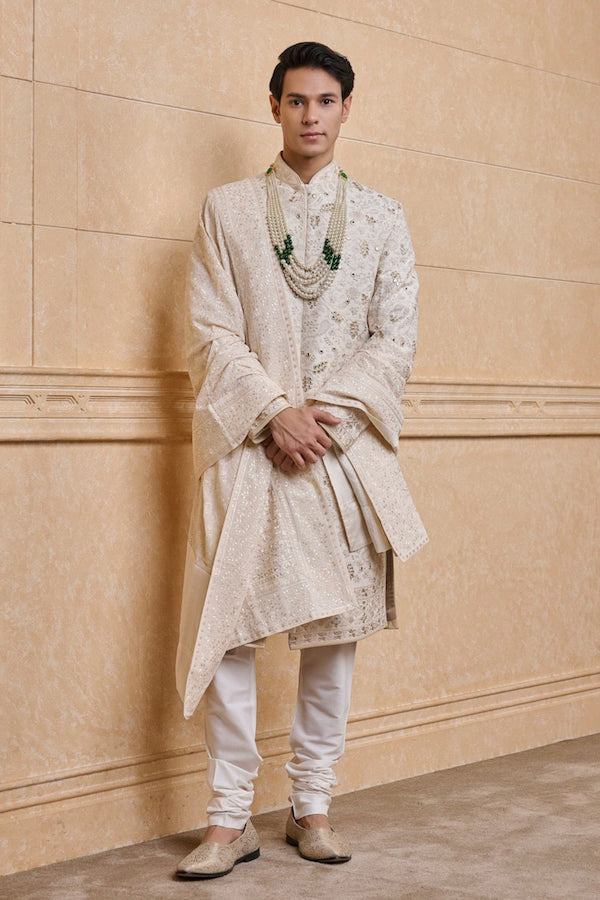 Man wearing designer engagement white dress for men with embroidered sherwani, layered stole, and pearl necklace, styled for a royal groom look